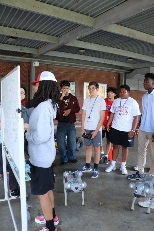 Alex Brown at a whiteboard with a group of students and robotic dogs at an NC State outreach event